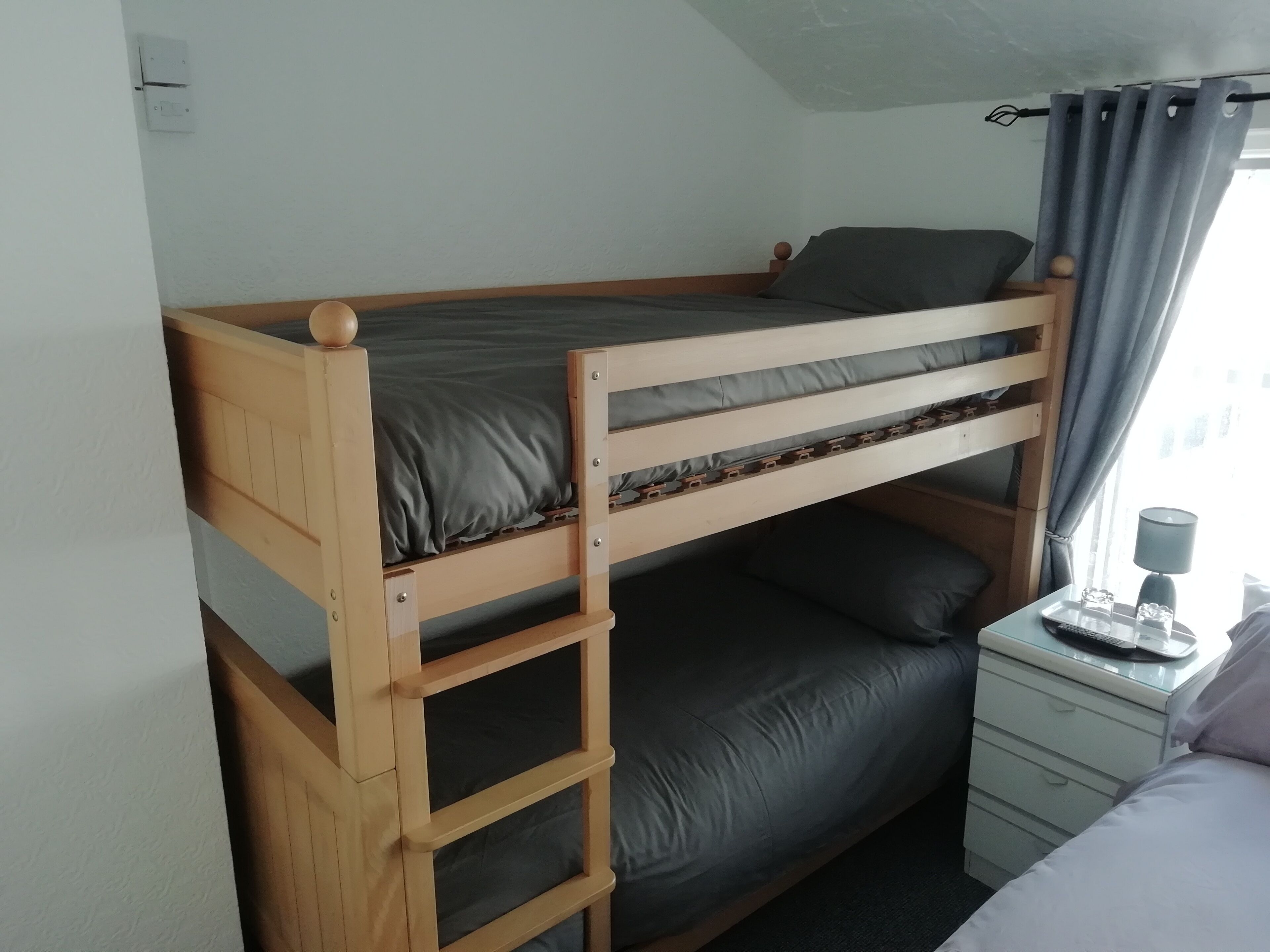 A bunk bed with a wooden frame and grey bedding, a bedside table with a lamp, and a window with curtains.