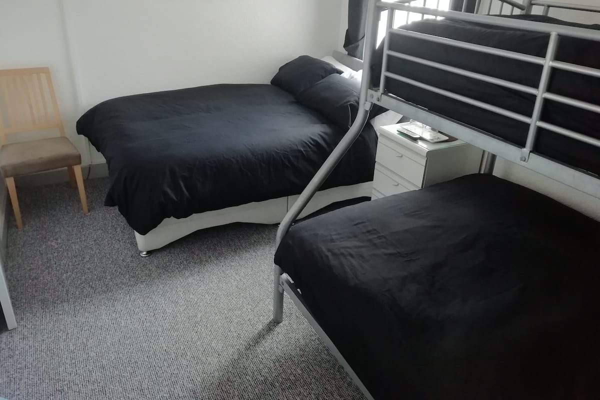 A compact room with a bunk bed, a single chair, a small bedside table with a remote control, and a wall-mounted picture.