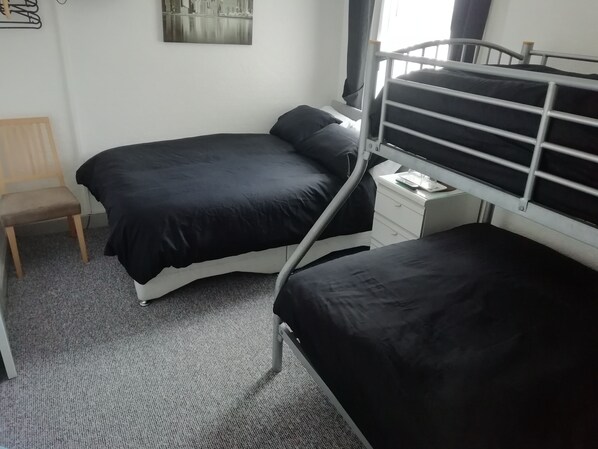 Habitación pequeña con una cama individual superpuesta, una silla, una mesita de noche con un control remoto y un cuadro colgado en la pared.
