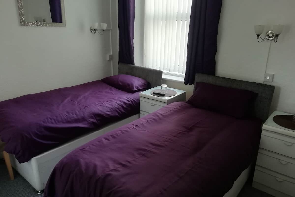 A bedroom with two single beds, each with a purple bedspread, a bedside table with a lamp, and a window with curtains.
