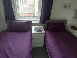 A twin bedroom with two beds, a small table with a remote control, and a window with curtains.