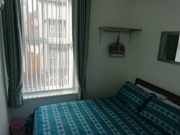 A bedroom with a bed, a window with blinds, a hanging light fixture, and a small shelf.