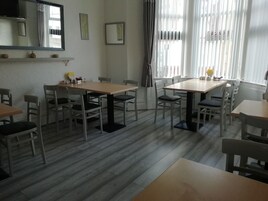 Een eetgedeelte met meerdere tafels en stoelen, een spiegel aan de muur en een raam met jaloezieën.