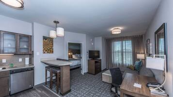 A compact hotel room with a kitchenette, a bed, a TV, a desk, and a seating area.