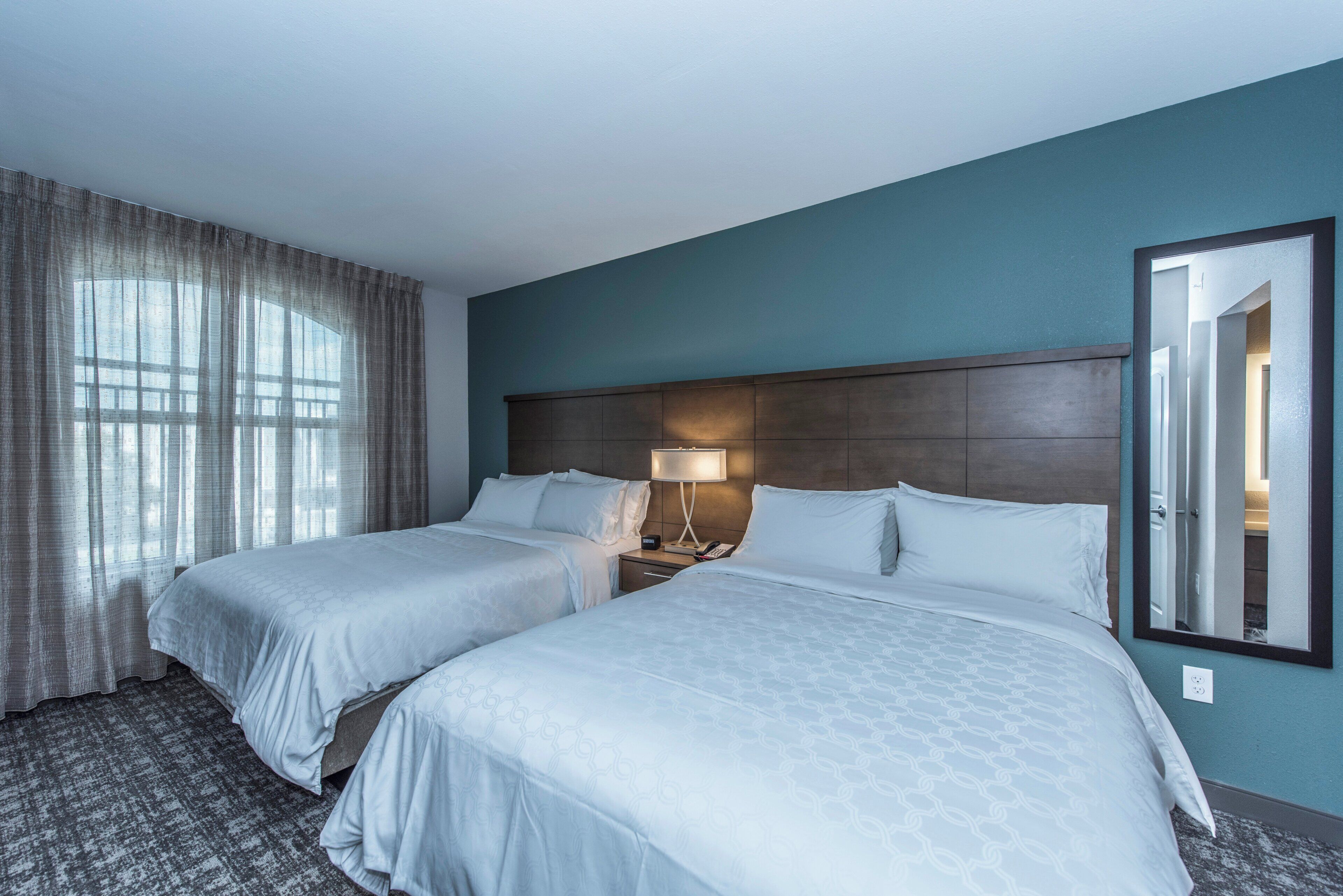 A hotel room with two beds, a nightstand with a lamp, a large mirror, and a window with curtains.