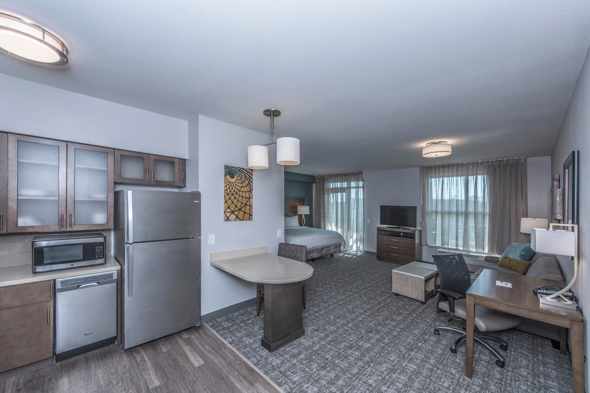 A compact hotel room with a kitchenette, a bed, a desk, and a TV.
