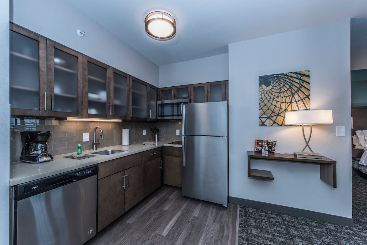 A modern kitchen with dark wooden cabinets, stainless steel appliances, a built-in microwave, and a refrigerator. There is a small shelf with a lamp and a wall art piece above the sink.