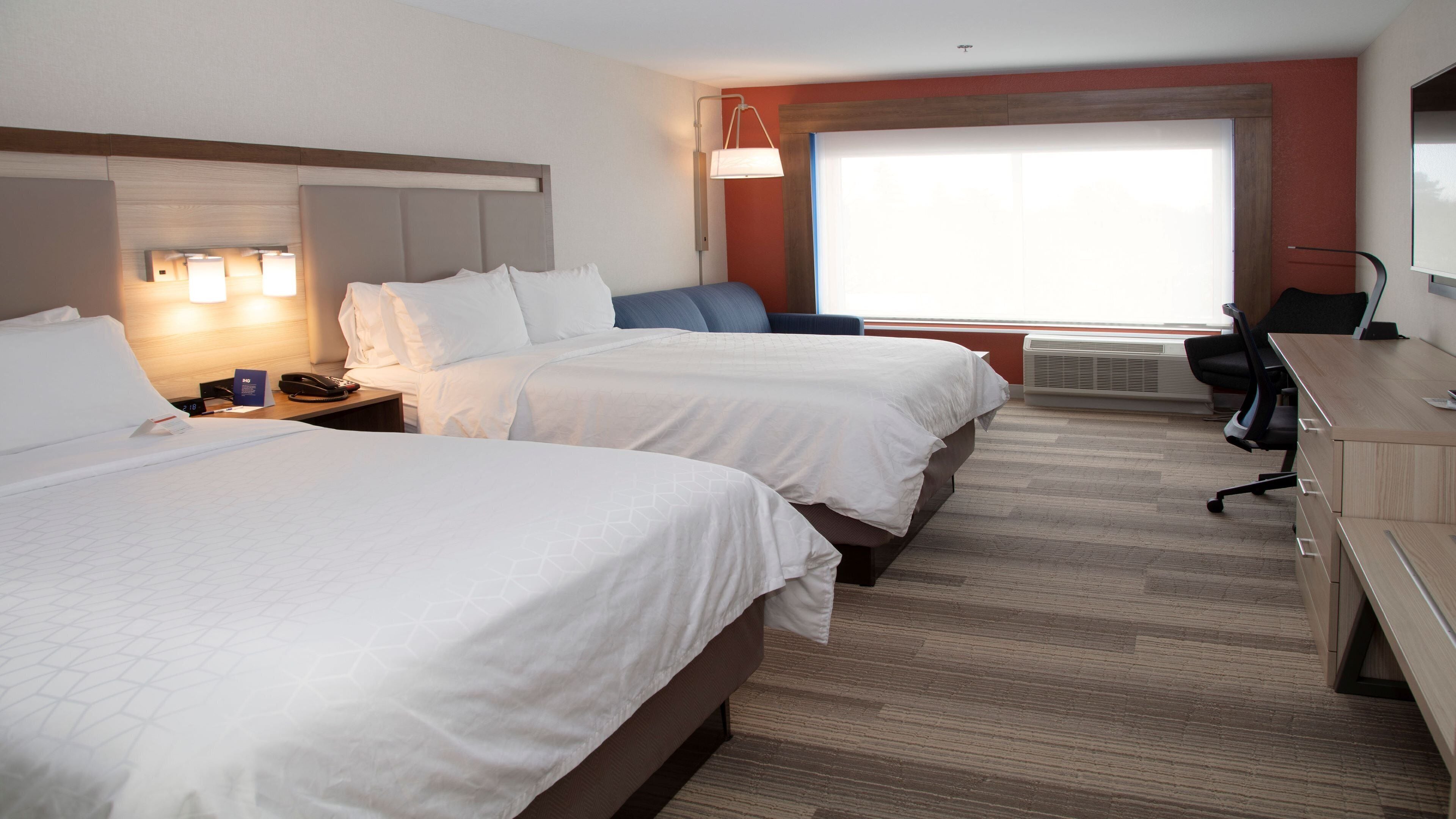 A hotel room with two beds, a desk, a chair, a lamp, and a large window.