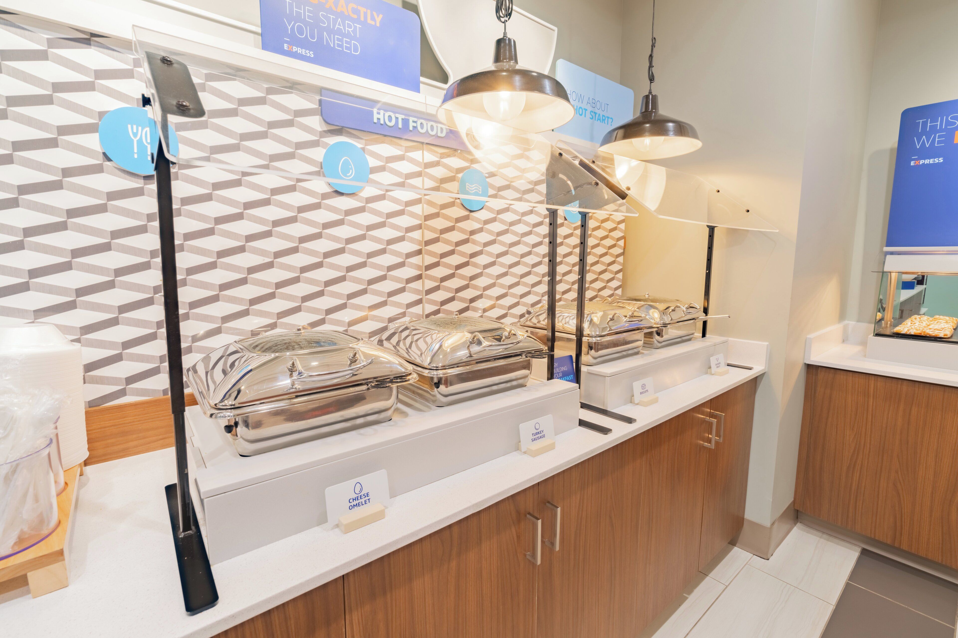 Un espace buffet avec des présentoirs de plats chauds clairement identifiés, un coin boissons en libre-service et un décor mural moderne aux motifs géométriques.
