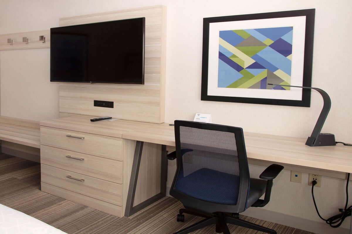 A hotel room with a wooden desk, a television, a framed abstract painting, and a black office chair.