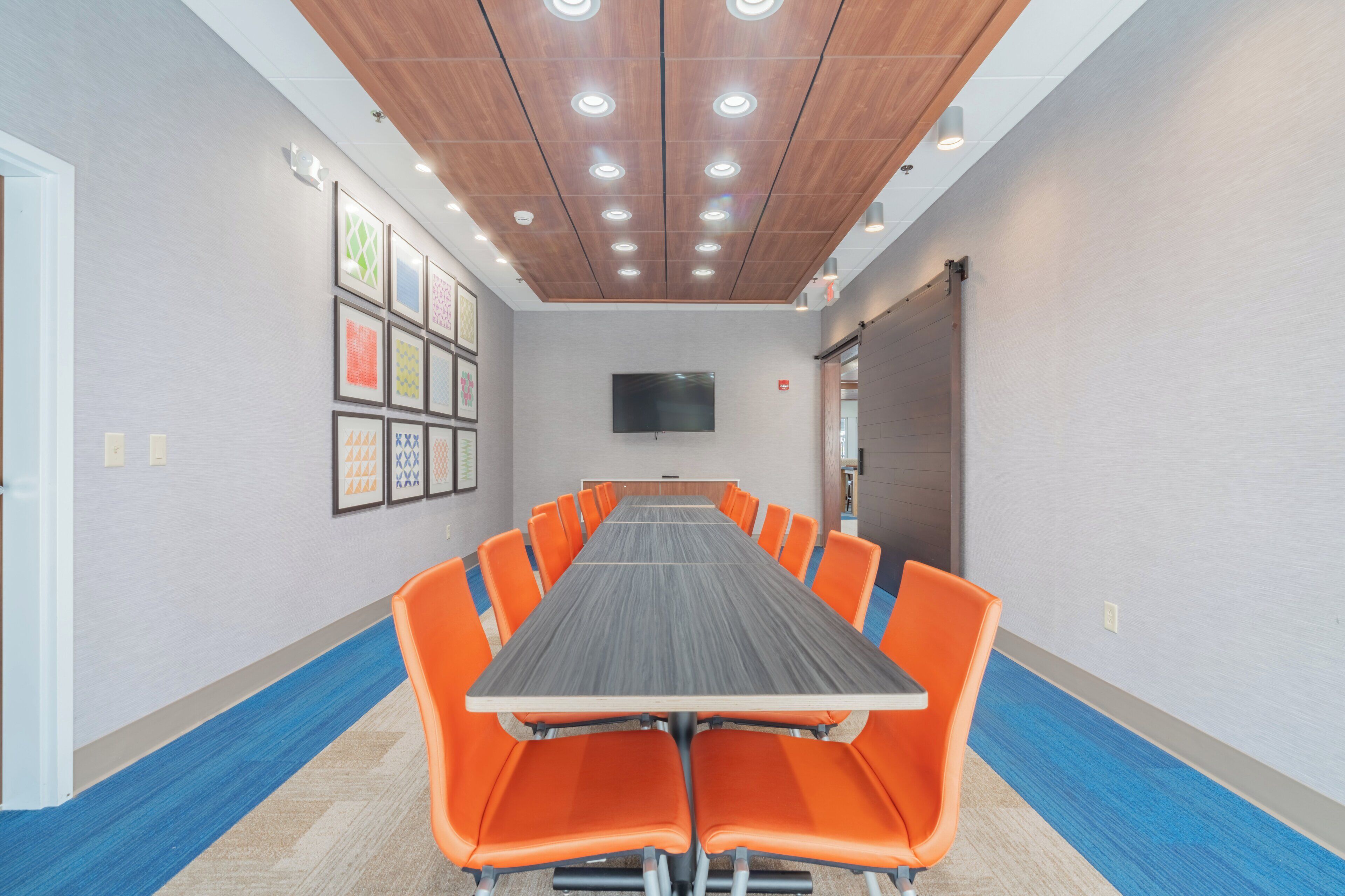 A conference room with a long table and orange chairs, a flat-screen TV on the wall, and a wooden ceiling with recessed lighting.