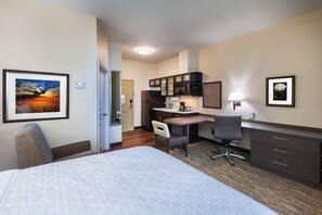 A hotel room with a bed, a desk, a chair, a kitchenette, and framed pictures on the walls.