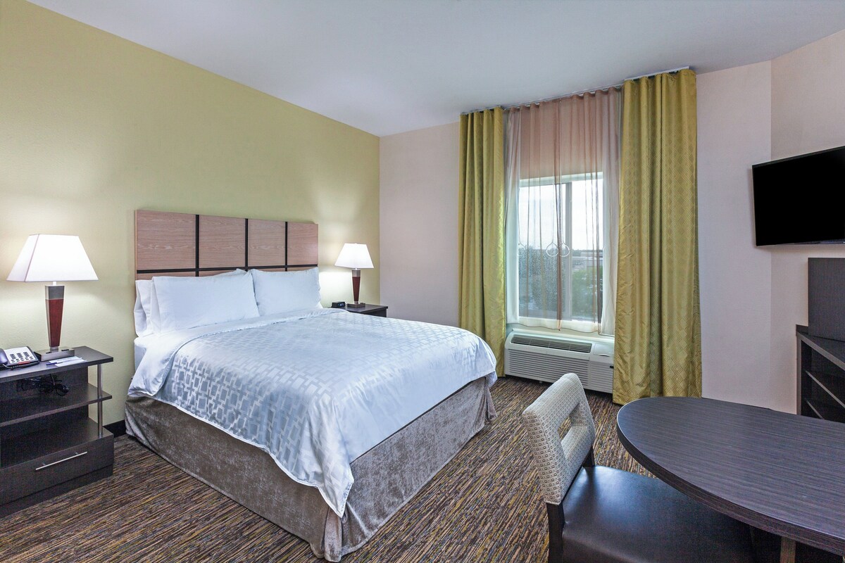 A hotel room with a bed, two bedside tables with lamps, a desk with a chair, a TV, and a window with curtains.