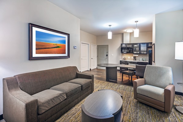 A modern hotel room with a sofa, armchair, and a round coffee table. There is a dining area with a table and chairs, and a kitchen area with cabinets and appliances. - Candlewood Suites Houston - Pasadena by IHG (Pasadena)