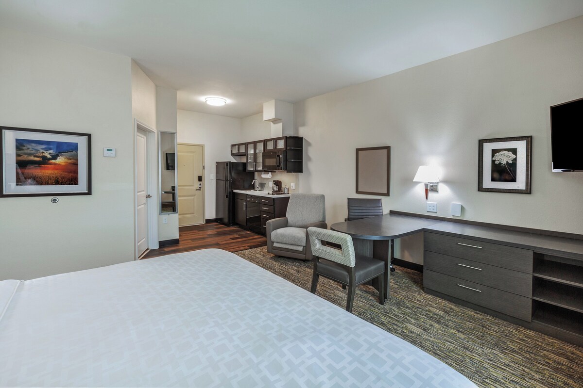 A hotel room with a bed, a desk, a chair, a kitchenette, and a television.