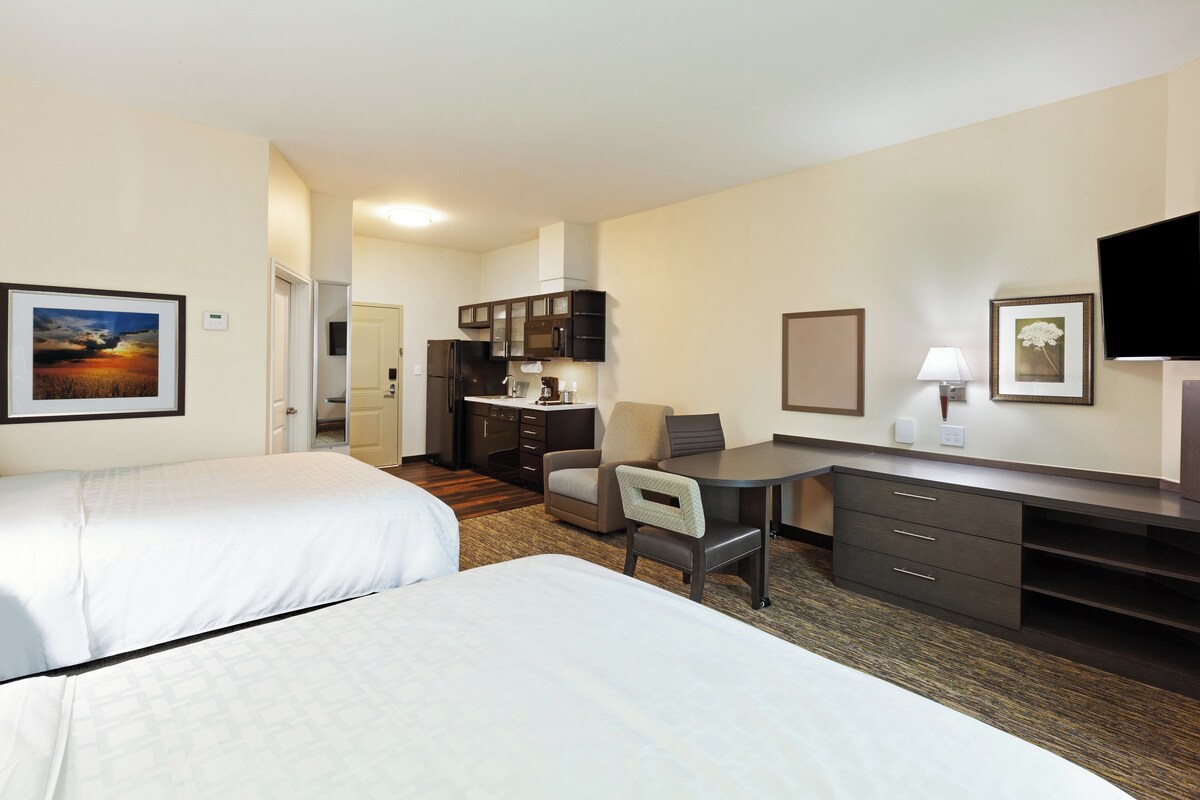 A hotel room with two beds, a desk, a chair, a TV, and a kitchenette.