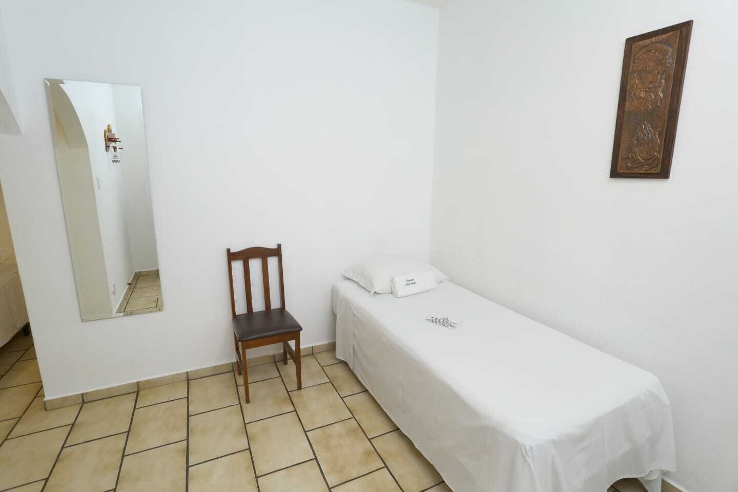 A small, clean room with a single bed, a chair, a bedside table, and a wall-mounted mirror.