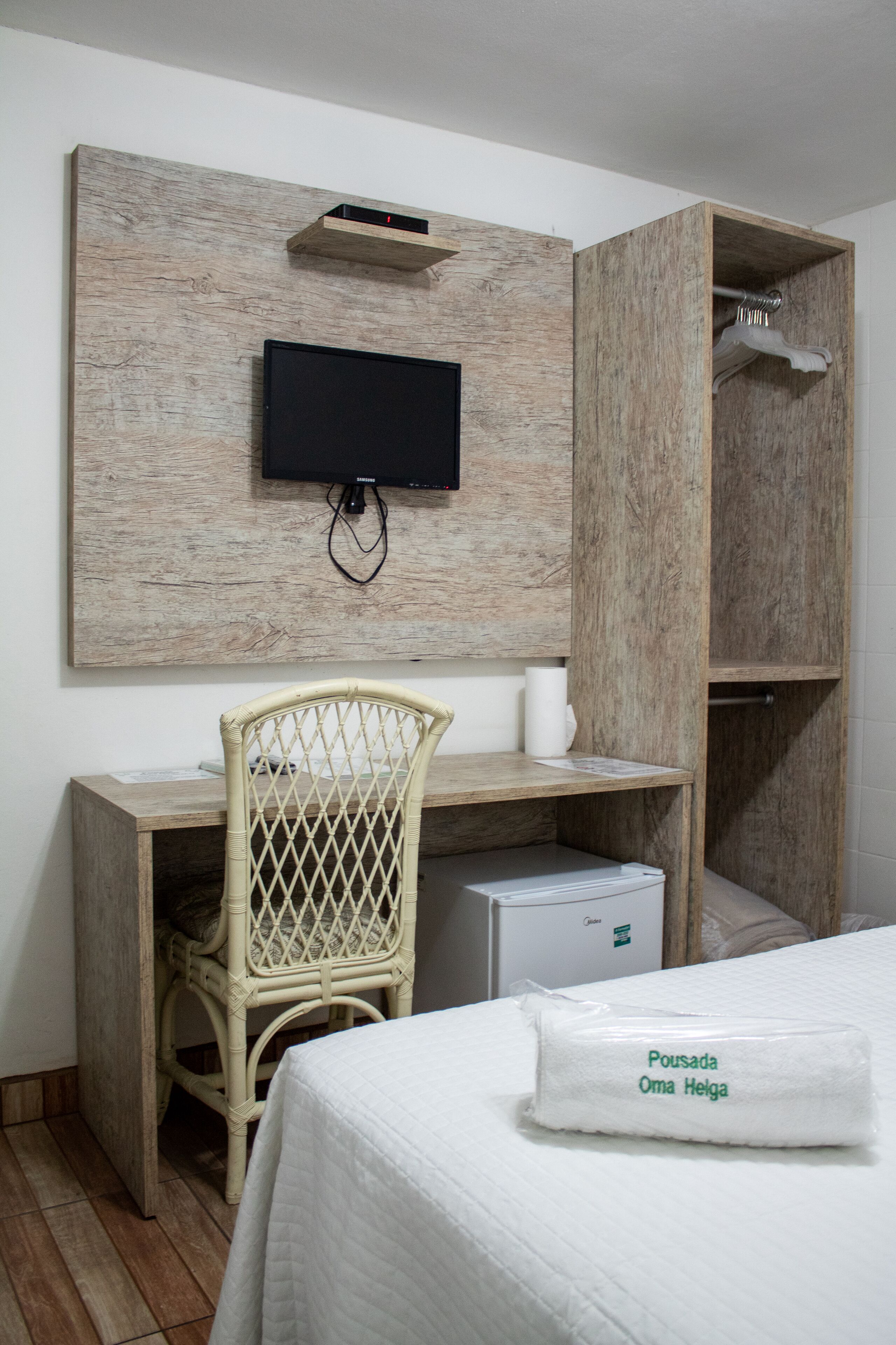 Una habitación de hotel con un escritorio de madera, un televisor montado en la pared, una silla de mimbre, una cama con edredón blanco y un refrigerador pequeño.