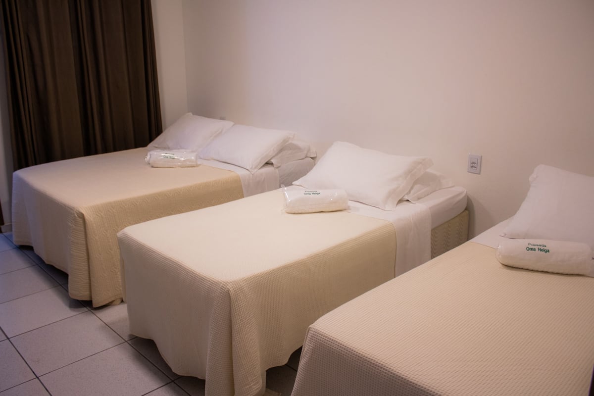 Tres camas individuales con ropa blanca y almohadas, piso de baldosas y una pared lisa con toma de corriente.
