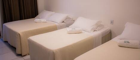 Tres camas individuales con ropa blanca y almohadas, piso de baldosas y una pared lisa con toma de corriente.