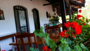 A patio area with wooden chairs and tables, a white wall with a window, and red flowers in the foreground.