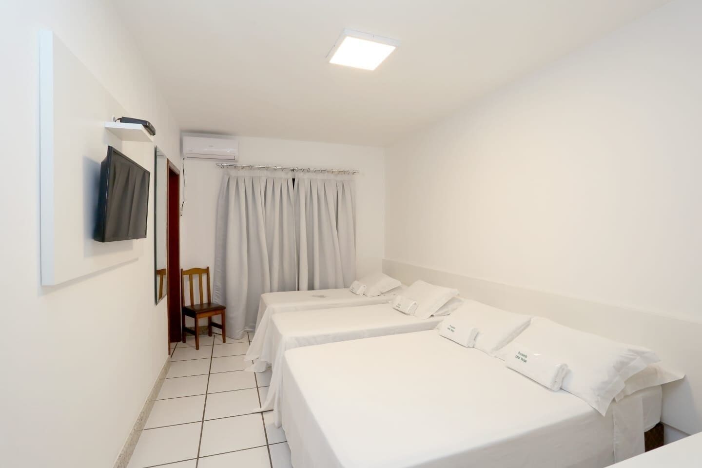 A hotel room with two single beds, a TV mounted on the wall, a chair, and an air conditioning unit.