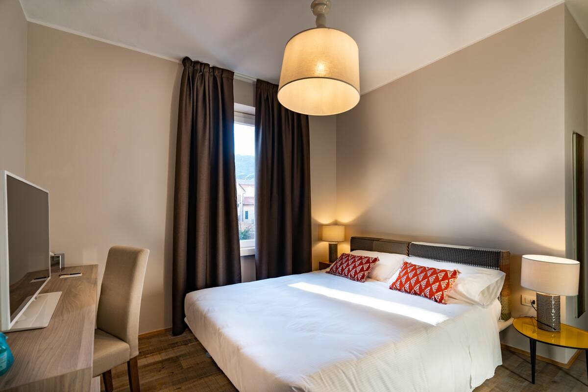 Une chambre d’hôtel avec un lit, un bureau, une chaise, une lampe et une fenêtre avec des rideaux.