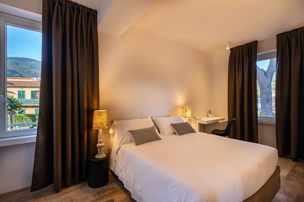 Une chambre d’hôtel avec un grand lit, deux tables de chevet avec des lampes, une chaise et une vue sur les bâtiments par la fenêtre.