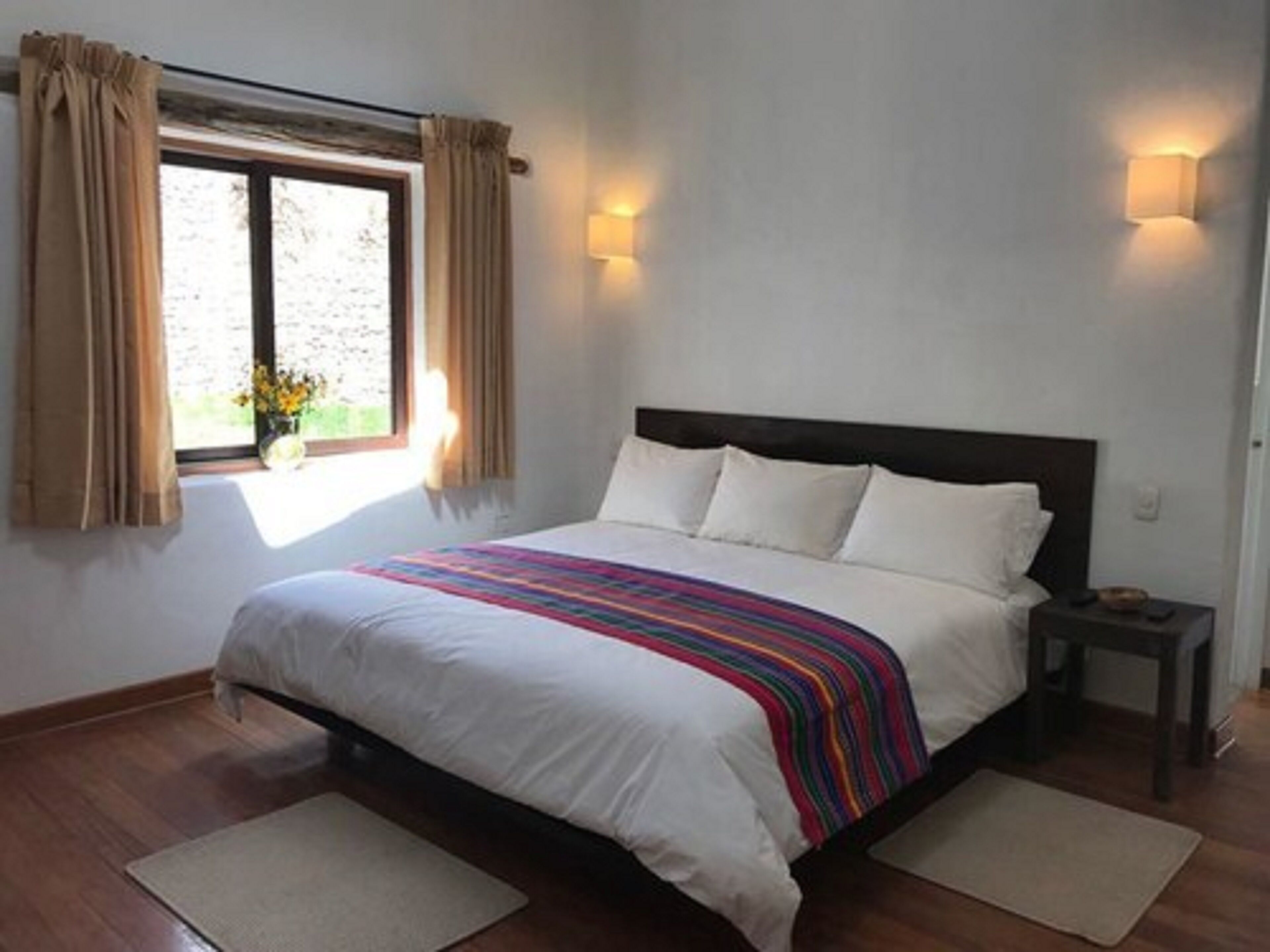 A bedroom with a bed, two bedside tables, a window with curtains, and a wall-mounted light fixture.