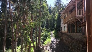 Una casa de madera con base de piedra y un balcón, rodeada de árboles altos y un sendero de tierra.