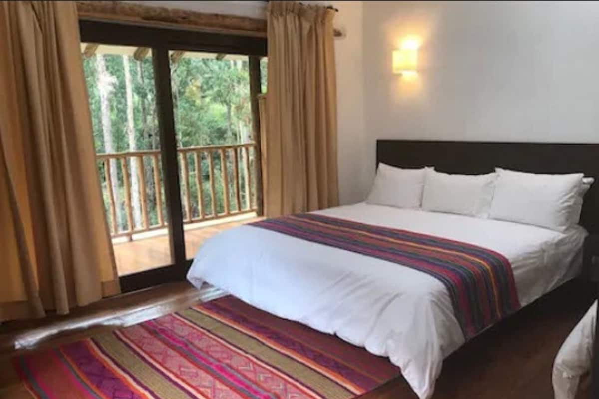 A bedroom with a bed, a window with curtains, a wooden floor, and a wall-mounted light.