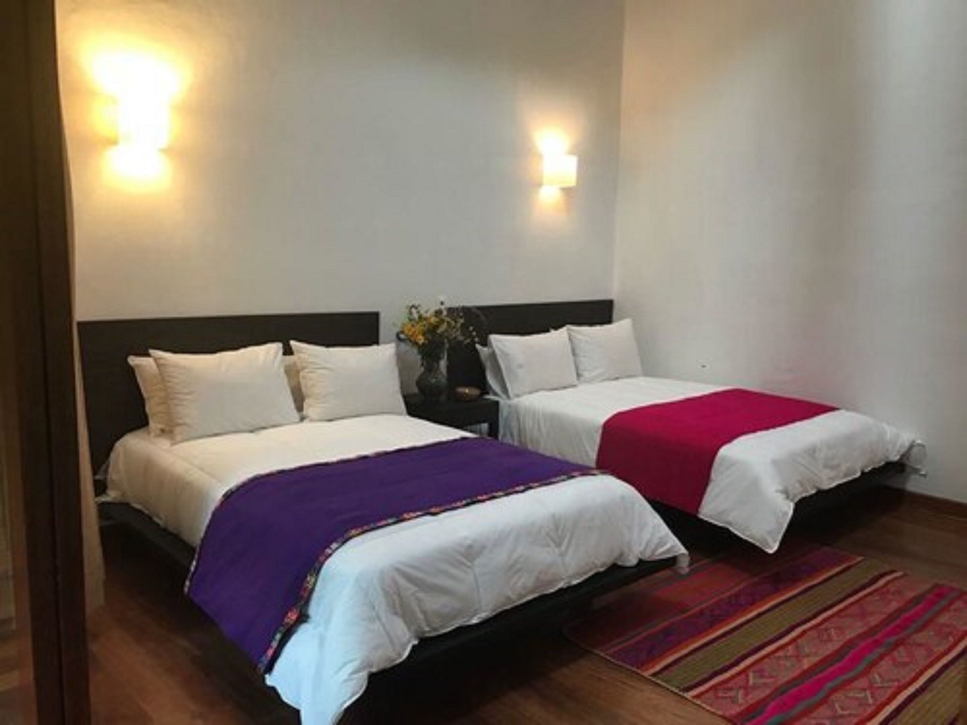 Habitación de hotel con dos camas, cada una con ropa blanca y una colcha morada y roja. Hay luces fijadas a la pared y piso de madera.