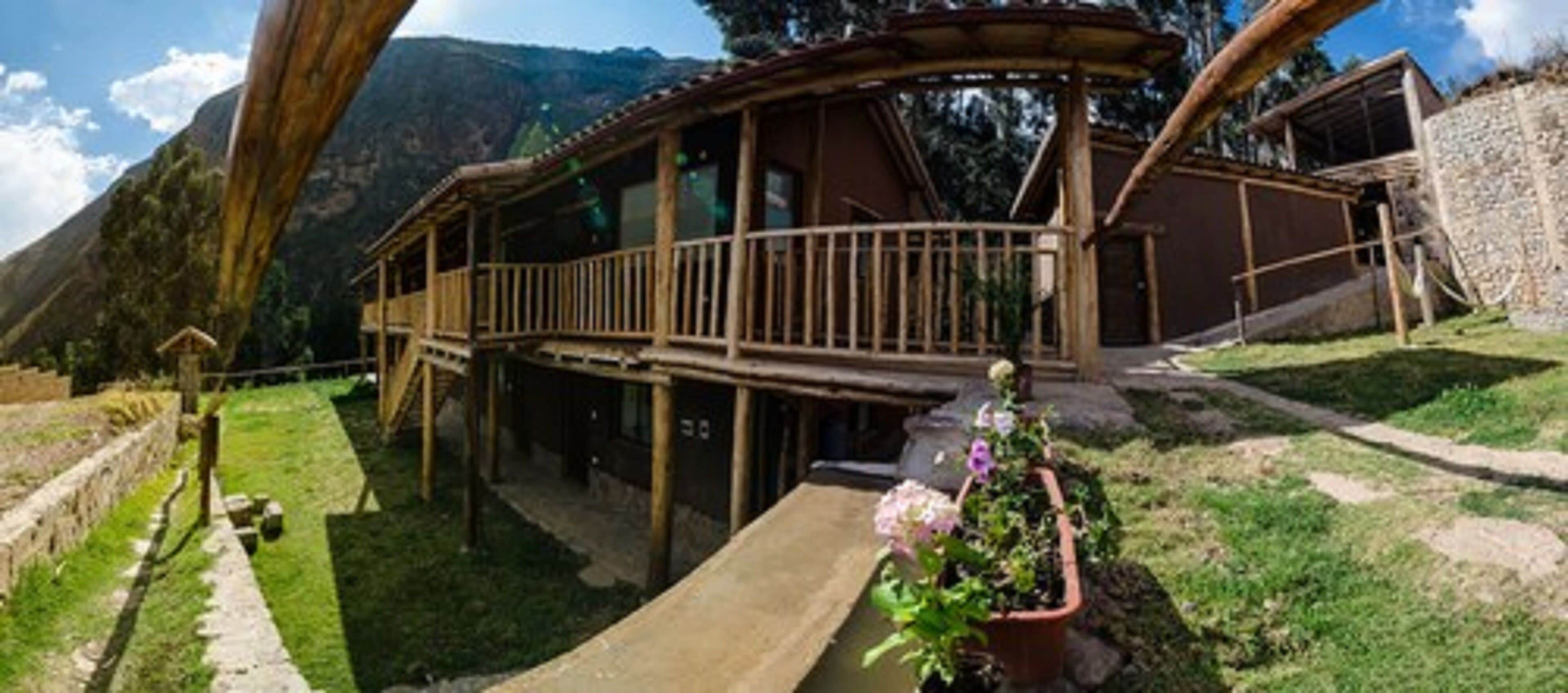 A wooden cabin with a balcony, surrounded by a stone wall and a grassy area with a flower pot.
