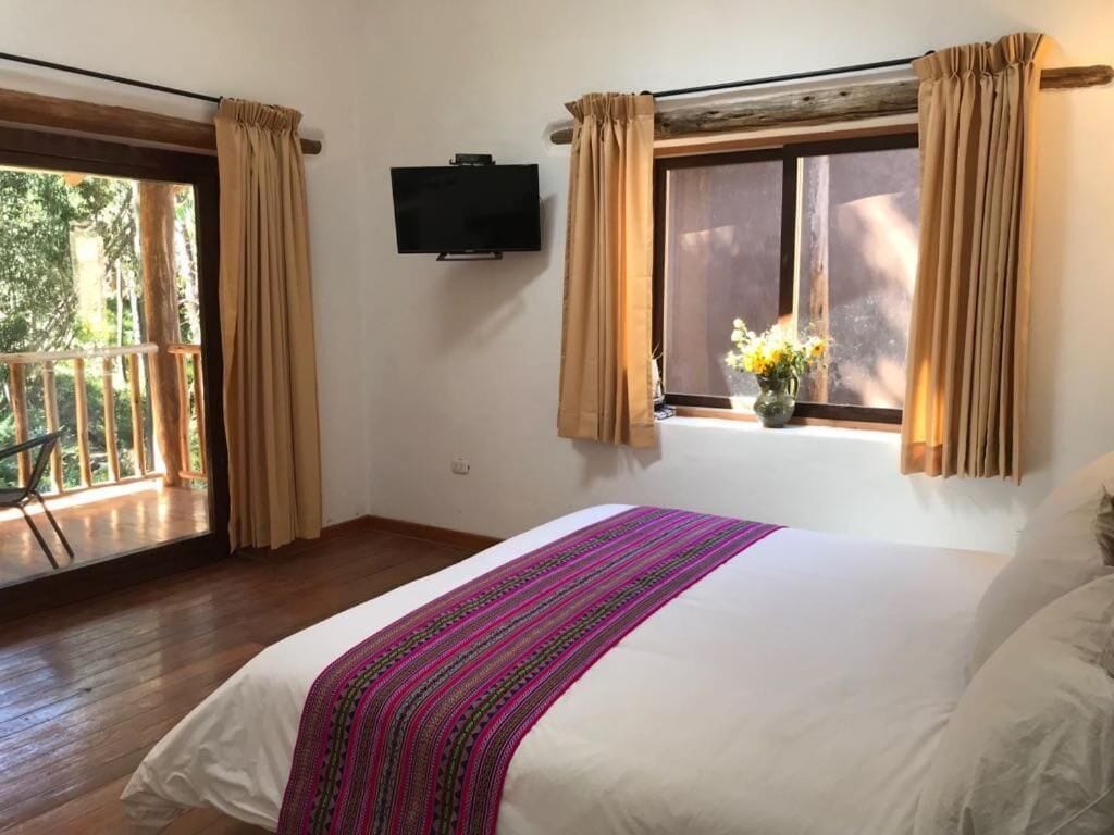 Un dormitorio con una cama grande, una ventana con cortinas, un televisor montado en la pared y un balcón con baranda de madera.
