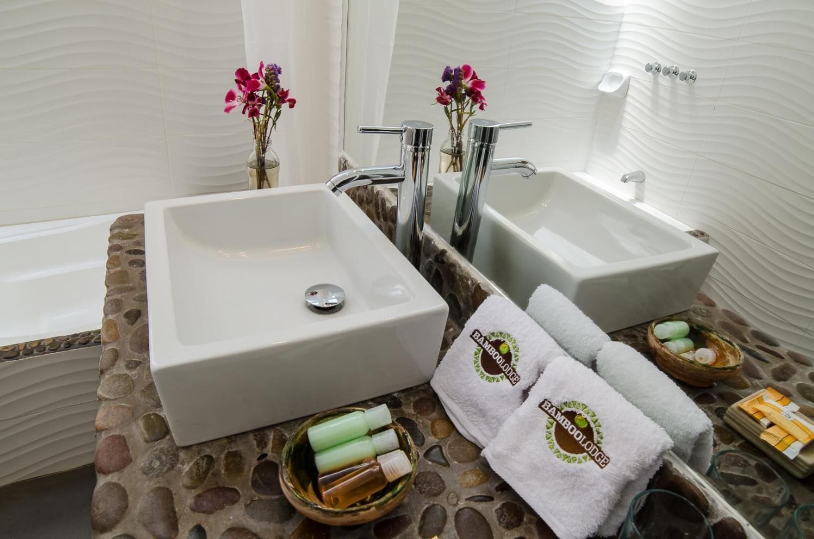 A bathroom with two sinks, a bathtub, and a stone-textured countertop. There are towels and toiletries on the counter.