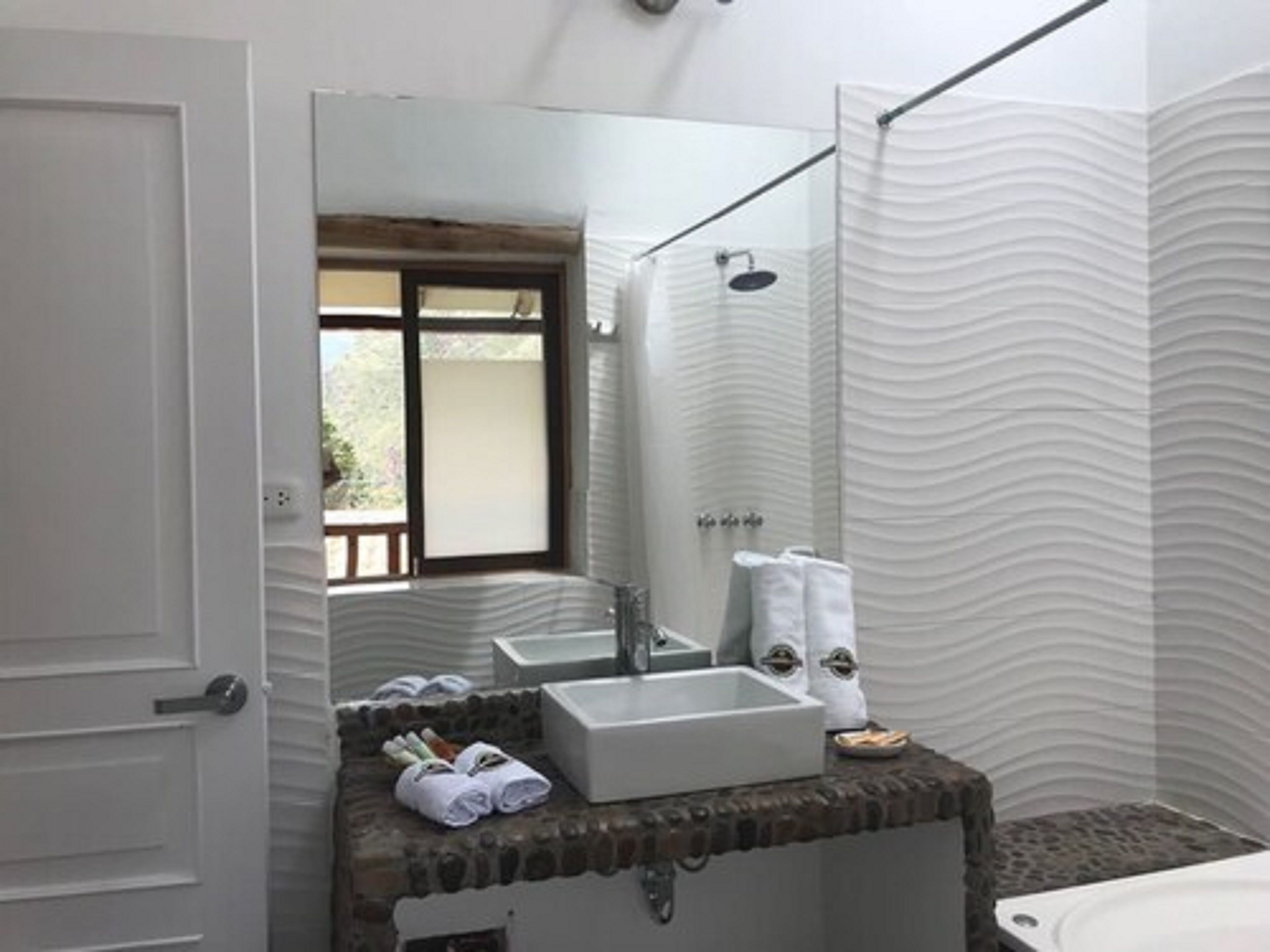 Un baño con un espejo grande, un lavamanos, una ducha y una bañera. Hay toallas y artículos de aseo en la encimera.