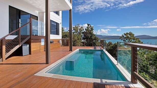 A modern outdoor pool area with a wooden deck, a glass balustrade, and a view of the sea and mountains. - Island Views - the perfect holiday home. Centre of Airlie Beach. (Airlie Beach)