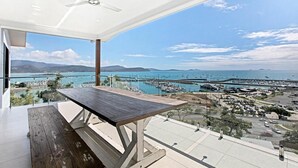 A balcony with a wooden table and a ceiling fan, overlooking a marina with boats and a coastal landscape. - Island Views - the perfect holiday home. Centre of Airlie Beach. (Airlie Beach)