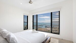 A bedroom with a large bed, two windows with black shutters, a ceiling fan, and a view of the sea and boats. - Island Views - the perfect holiday home. Centre of Airlie Beach. (Airlie Beach)