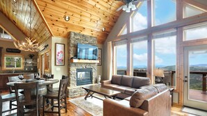 A spacious living room with a stone fireplace, a large TV, and a view of the mountains through large windows.
