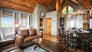 A spacious living room with a brown leather sofa, a wooden dining table with chairs, and a large window with curtains.