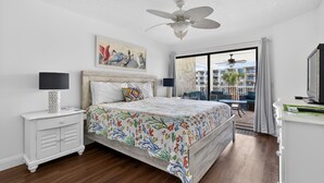 A bedroom with a bed, a ceiling fan, a nightstand, a lamp, a painting, and a view of the outside. - CRC 2208 - Oceanview Condo (St Augustine)