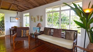 Un salon chaleureux avec un canapé en bois, une table à manger et des chaises. De grandes fenêtres offrent une vue sur la verdure environnante.