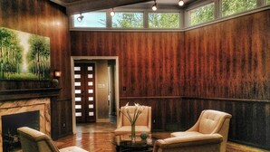 Un hall d’entrée spacieux avec une cheminée, deux fauteuils, une table basse en verre et un tableau représentant une forêt.