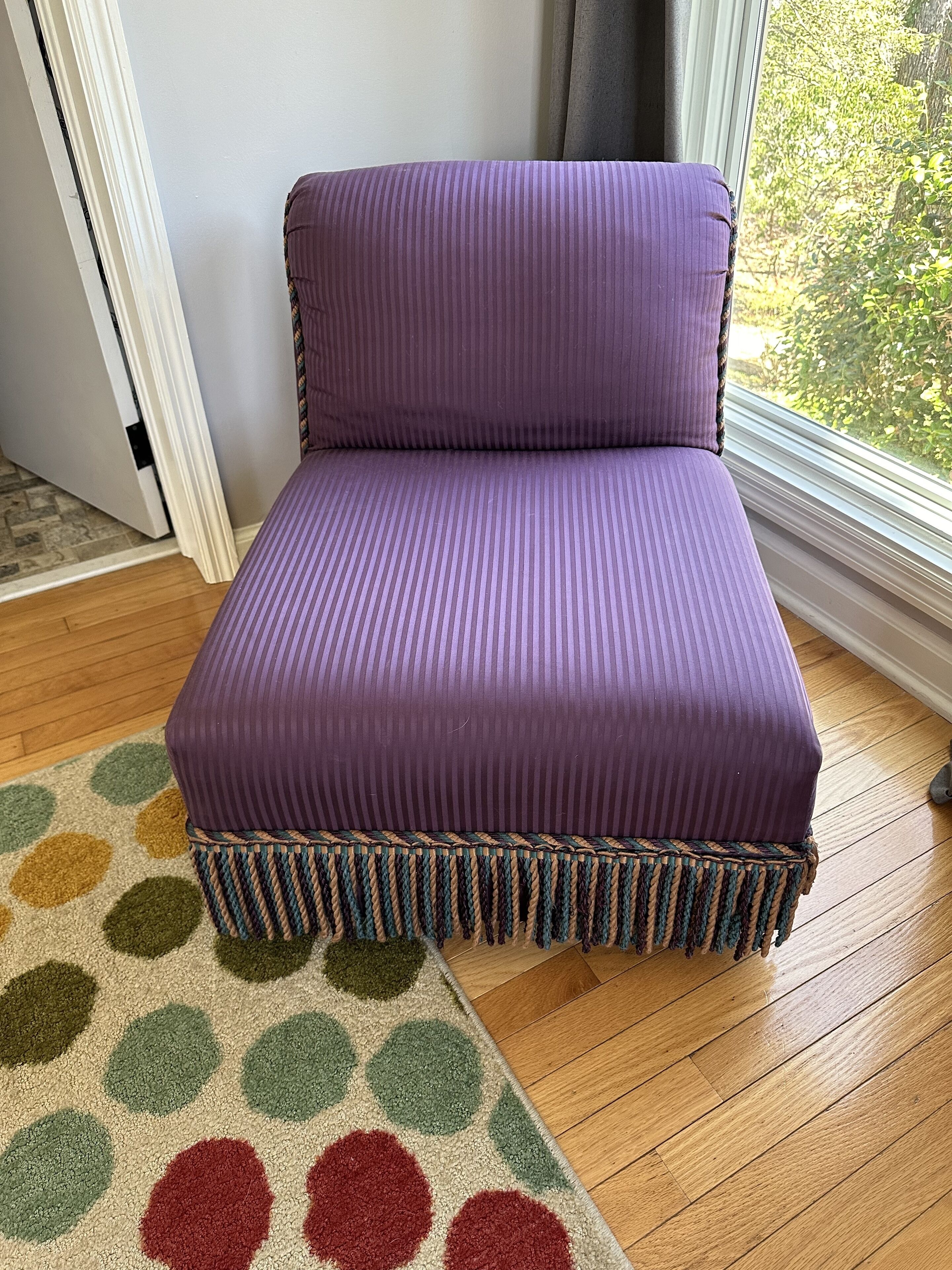 A purple sofa with a braided base, positioned on a wooden floor next to a window with a view of greenery.