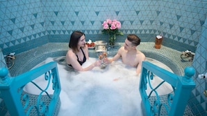 Una pareja en una bañera de hidromasaje turquesa con una pared de azulejos geométricos, un jarrón de rosas rosadas y una copa de champán.