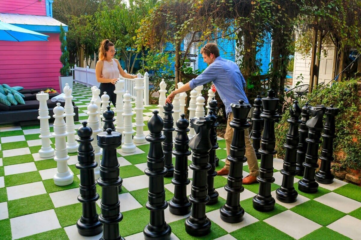 A large outdoor chessboard with oversized chess pieces, set against a backdrop of greenery and a pink building.
