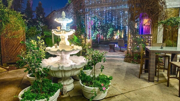 Un jardín con una fuente central, plantas en macetas y un área de descanso con mesas y sillas.