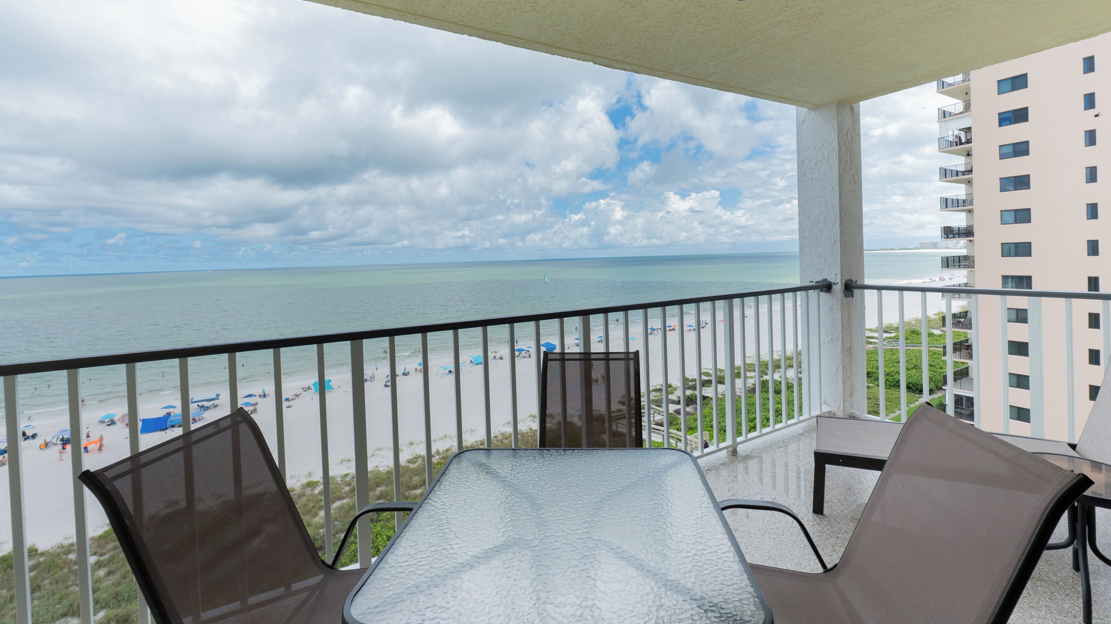 Un balcon avec une table en verre et deux chaises, donnant sur une plage parsemée de parasols, avec vue sur l’océan et un bâtiment situé de l’autre côté de l’eau.