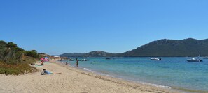 A beach with clear water, boats, and mountains in the background.
